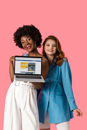 KYIV, UKRAINE - AUGUST 9, 2019: cheerful multicultural girls smiling near laptop with booking website on screen isolated on pinkのeditorial素材