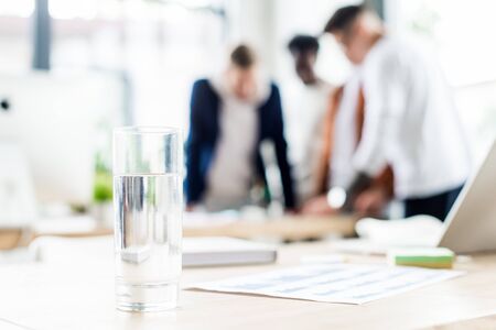 selective focus of glass with water on desk near businesspeople standing at workplace in officeの写真素材