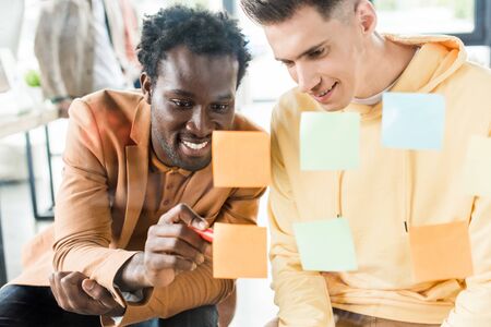 smiling african american businessman writing on sticky note while sitting near colleague in office