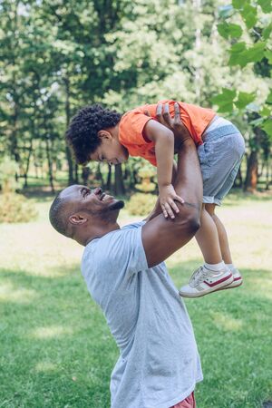 happy african american man holding son above head while walking in parkの写真素材