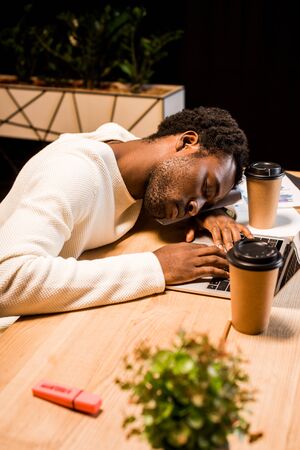tired african american businessman sleeping at workplace near laptop and disposable cups at night in officeの写真素材