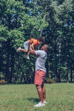 happy african american man holding cheerful son above head while having fun in parkの写真素材
