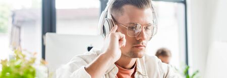 panoramic shot of thoughtful businessman sitting in headphones and using laptop in officeの写真素材