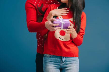 cropped view of couple in christmas sweaters holding surprise present isolated on blue