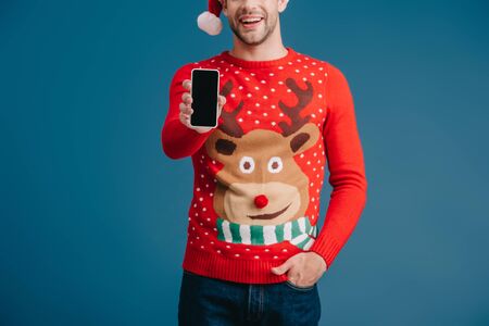 cropped view of man in santa hat and christmas sweater showing smartphone with blank screen, isolated on blueの写真素材