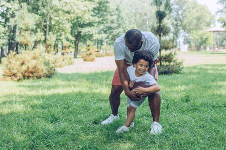 cheerful african american boy playing rugby with father in parkの写真素材
