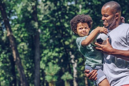 cute african american boy holding soccer ball while sitting on fathers handsの写真素材