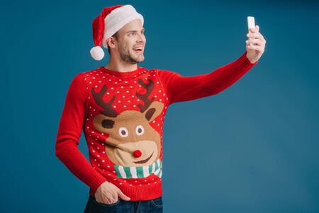 handsome happy man in santa hat and christmas sweater taking selfie on smartphone, isolated on blueの写真素材