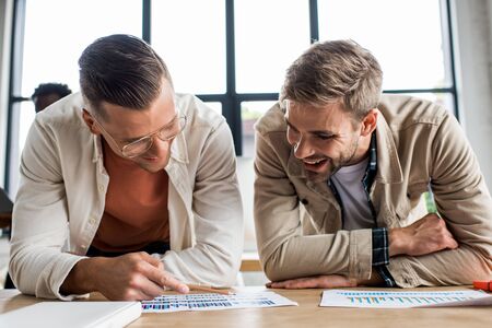 two young smiling businessmen analyzing papers with graphs and charts while working on startup...