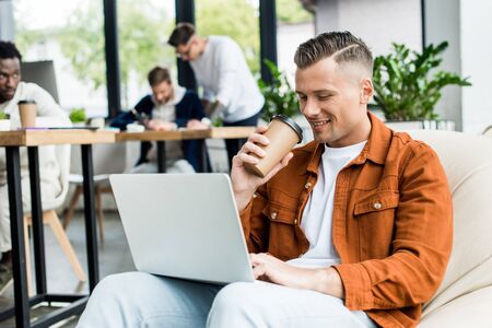 young businessman drinking coffee to go and using laptop while working in office near multicultural colleaguesの写真素材