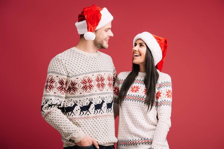 beautiful couple posing in santa hats and sweaters isolated on red