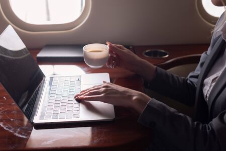 cropped view of businesswoman holding coffee cup and working on laptop in plane during business trip