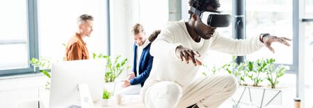 panoramic shot of african american businessman using vr headset and squatting on desk while colleagues working in officeの写真素材