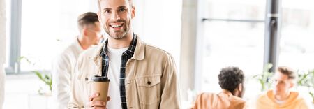 panoramic shot of young businessman holding coffee to go and smiling at camera