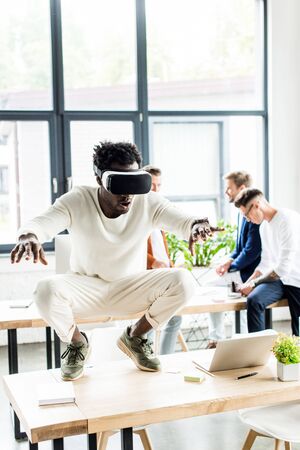 african american businessman squatting on table while using vr headset, and colleagues working in...