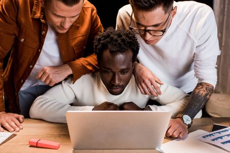 attentive multicultural businesspeople looking at laptop while working at night in officeの写真素材