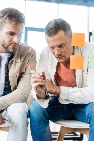 young businessman writing on glass board while working with colleague in office