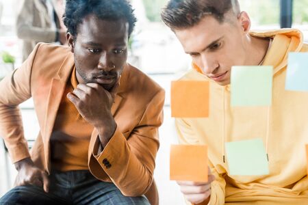 two young multicultural businessmen looking at sticky notes fixed on glass board in officeの写真素材