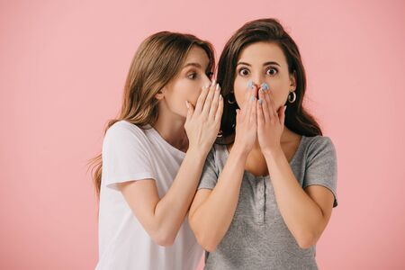 attractive woman telling secret to her shocked friend in t-shirt isolated on pink