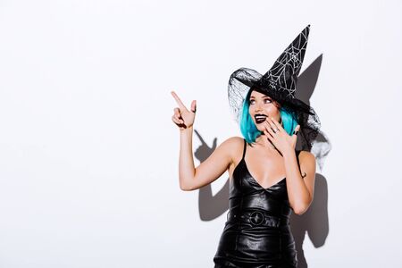 smiling girl in black witch Halloween costume with blue hair pointing with finger on white...