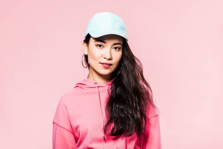 attractive asian woman in pink pullover and cap looking at camera isolated on pinkの写真素材
