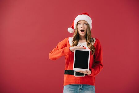 attractive woman in santa hat and sweater holding digital tablet isolated on redの写真素材