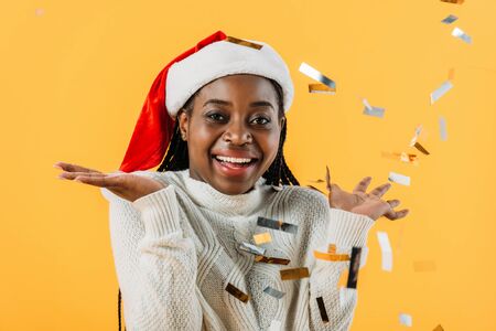 happy African American woman in Santa hat smiling and looking at camera on yellow background with confettiの写真素材