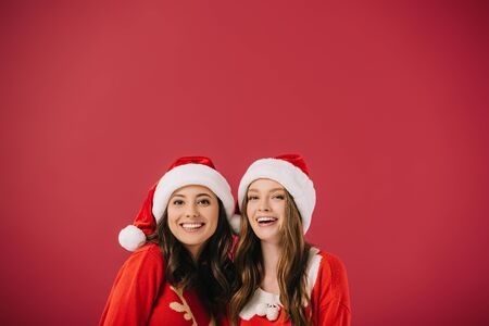 attractive and smiling women in sweaters and santa hats looking at camera isolated on redの写真素材