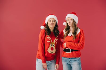 attractive and smiling women in sweaters and santa hats making faces isolated on redの写真素材