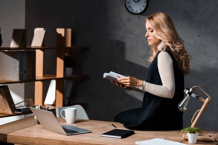 attractive and blonde businesswoman holding papers and sitting on table in office