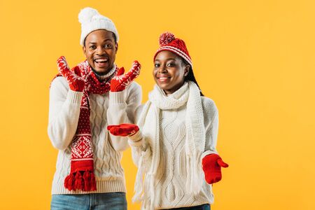 African American couple in knitted sweaters, scarfs and gloves smiling and looking at camera isolated on yellowの写真素材