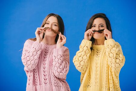 attractive women in sweaters playing with hair looking at camera isolated on blue