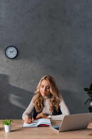 attractive and blonde businesswoman sitting at table and writing in notebookの写真素材
