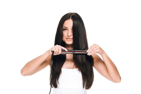 brunette beautiful woman straightening long shiny hair with flat iron isolated on white