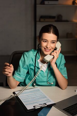 smiling nurse in uniform sitting at table and talking on telephone during night shiftの写真素材