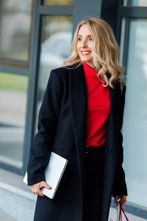 attractive businesswoman in black coat smiling and holding laptopの写真素材