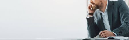 panoramic shot of businessman in formal wear talking on smartphone and holding penの写真素材