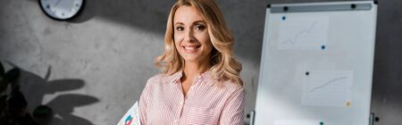 panoramic shot of attractive and blonde businesswoman smiling and looking at camera