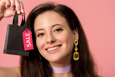 happy stylish disco girl holding shopping bag with sale lettering on label isolated on pink