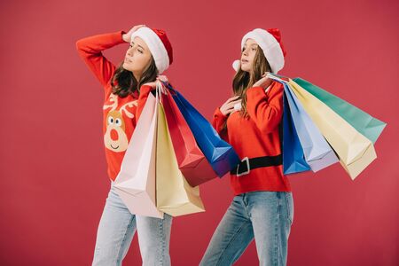 attractive women in sweaters and santa hats holding shopping bags and looking away isolated on redの写真素材