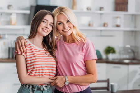 happy mother and daughter hugging and holding hands while smiling at camera in kitchenの写真素材