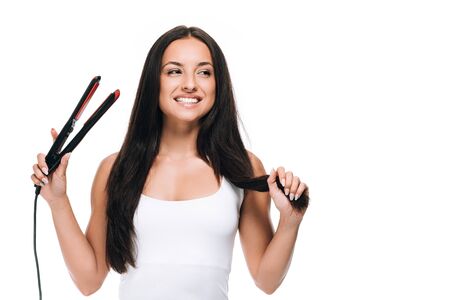 happy brunette beautiful woman straightening long shiny hair with flat iron isolated on whiteの写真素材