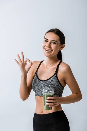smiling sportswoman holding smoothie and showing okay sign isolated on greyの写真素材