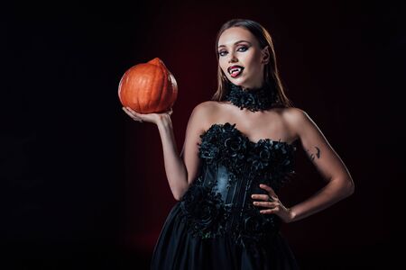 scary vampire girl with fangs in black gothic dress holding Halloween pumpkin on black background