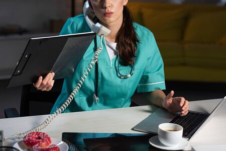 cropped view of nurse in uniform talking on telephone and holding clipboard during night shiftの写真素材