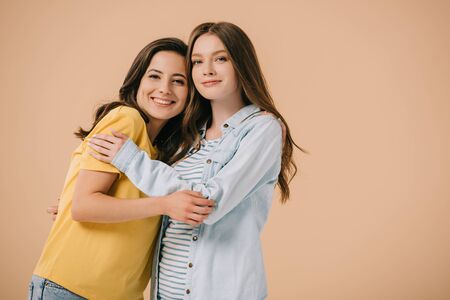 attractive and smiling friends hugging and looking at camera isolated on beige