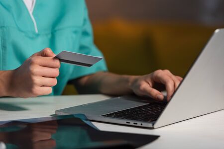 cropped view of nurse in uniform holding credit card and using laptop during night shift