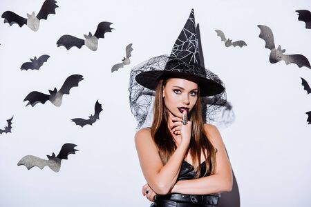 girl in black witch Halloween costume looking at camera near white wall with decorative batsの写真素材