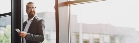 panoramic shot of handsome businessman in formal wear and glasses holding smartphoneの写真素材