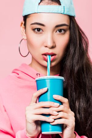 attractive asian woman in pullover and cap drinking from plastic cup isolated on pink